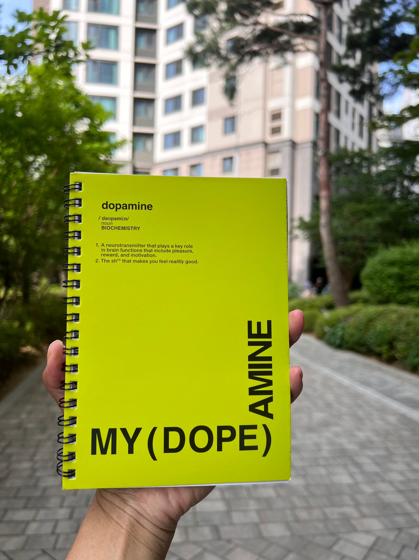 Yellow notebook titled 'MY (DOPE) AMINE' held in front of a blurred outdoor background with trees and buildings.