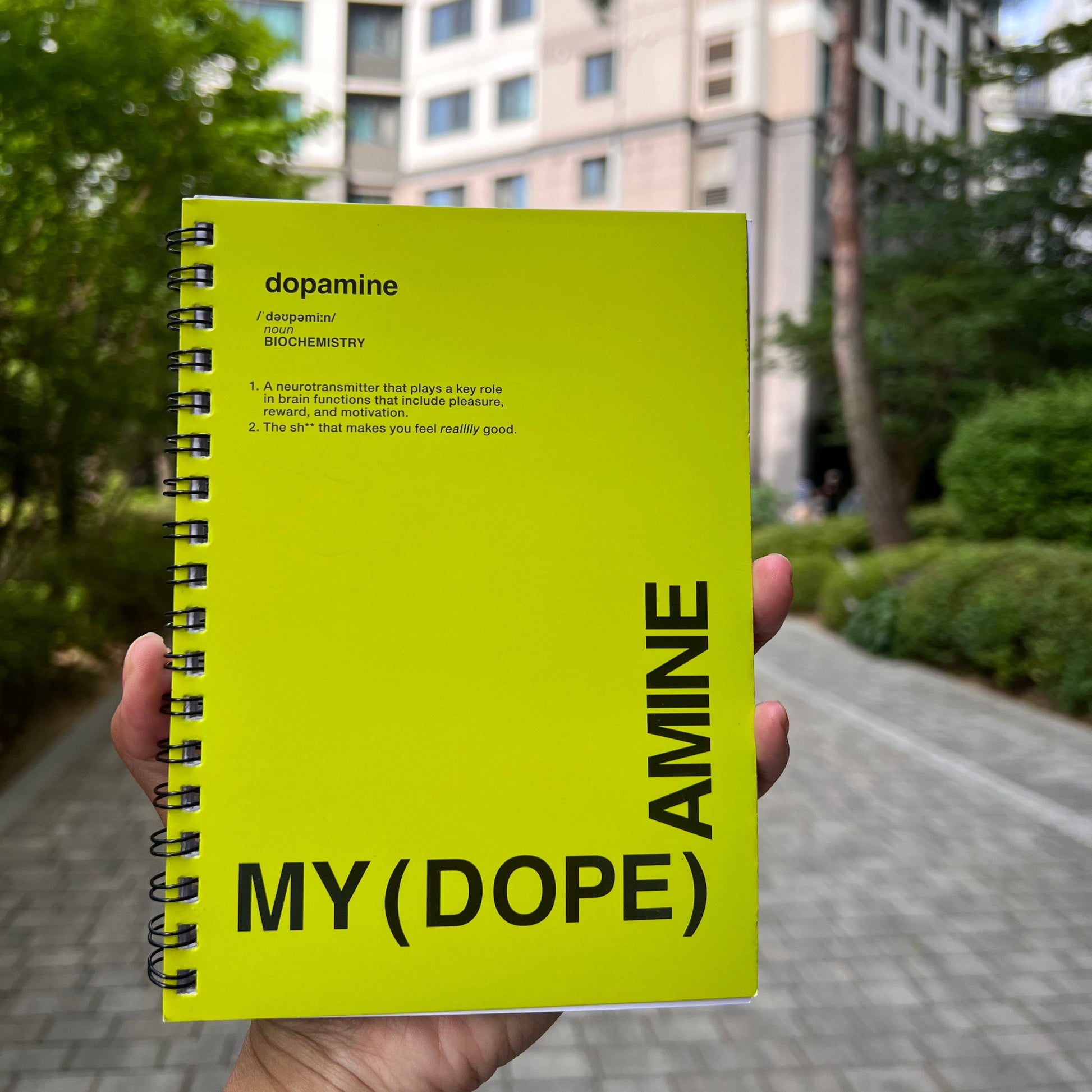 Yellow notebook titled 'MY (DOPE) AMINE' held in front of a blurred outdoor background with trees and buildings.