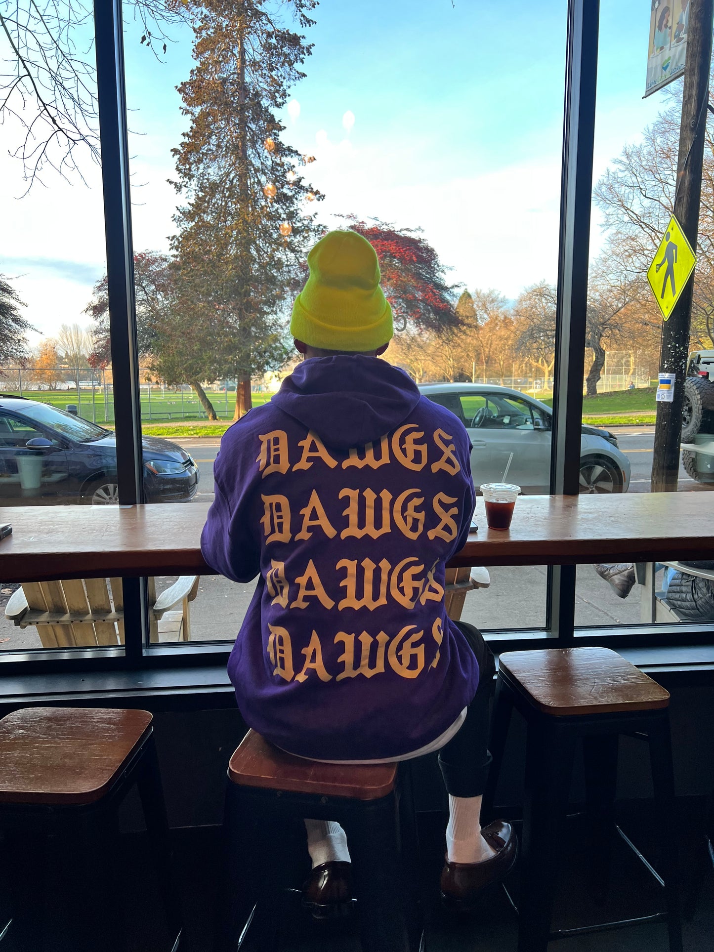 Person wearing a purple sweatshirt with 'DAMN' printed on it, sitting inside a cafe in front of a window