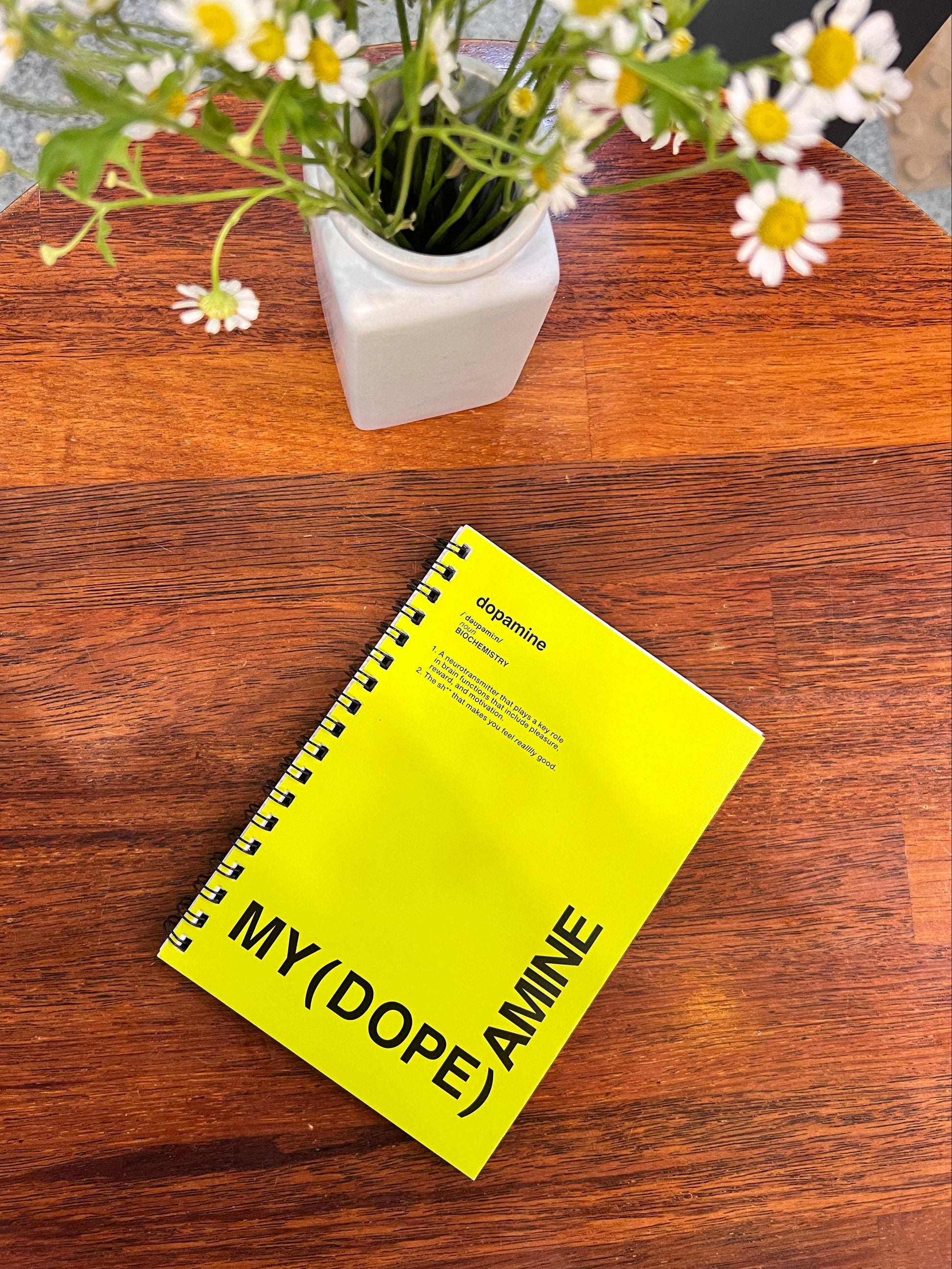Yellow notebook with text on a wooden surface next to a vase of flowers